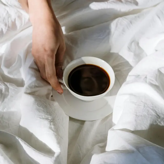 A close-up of a person's hands holding a white cup of coffee, sitting on a white bed sheet.