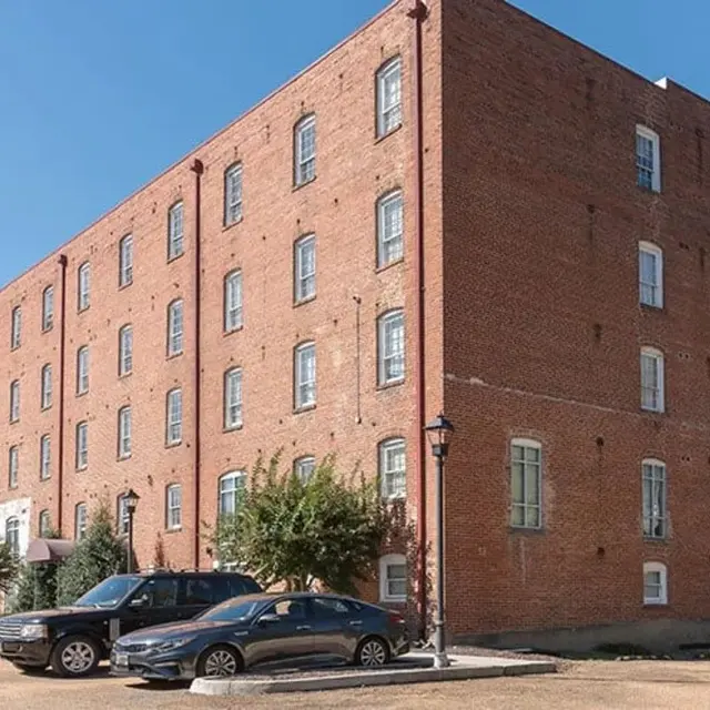 A historic red brick building with multiple windows, surrounded by cars and greenery, under a clear blue sky.