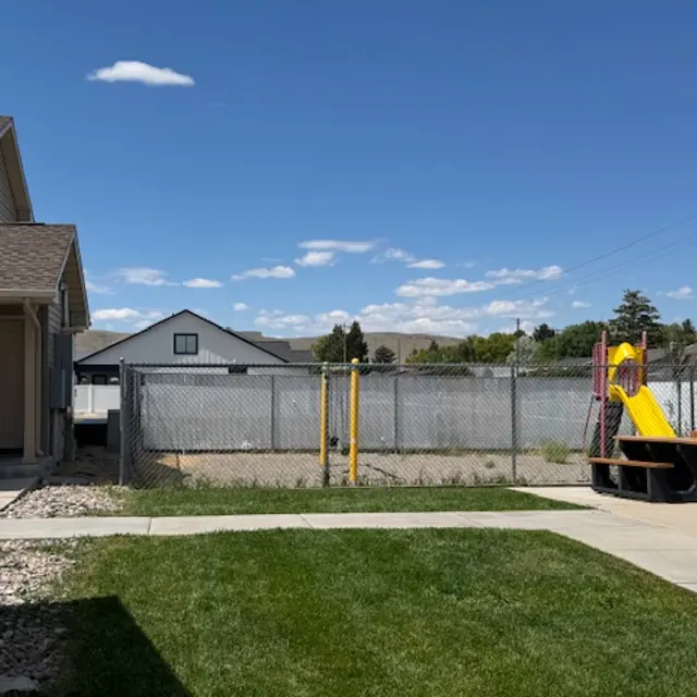 A view of a backyard featuring a playground and a fenced area with grass, a pathway, and buildings in the background under a blue sky with clouds.