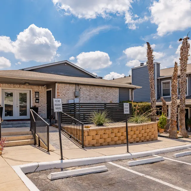 Exterior view of an apartment complex featuring a brick building, landscaped area, and parked cars under a blue sky with clouds.