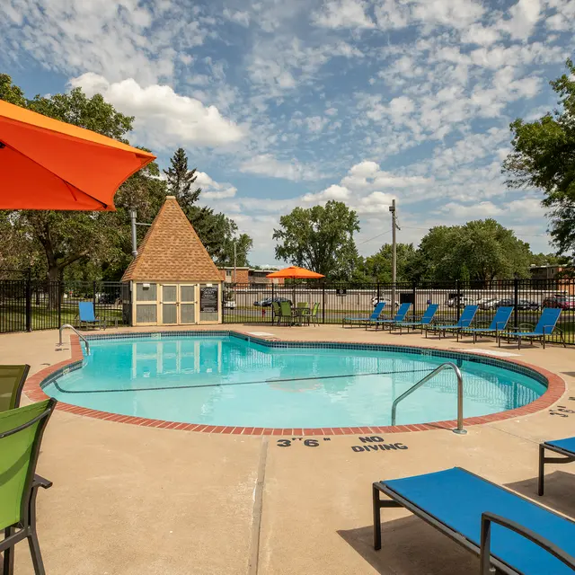 Regency Park - A vibrant pool area with a clear blue swimming pool surrounded by lounge chairs, orange umbrellas, and green seating. A small, triangular-roofed structure is located nearby, with trees and a partially cloudy sky in the background.