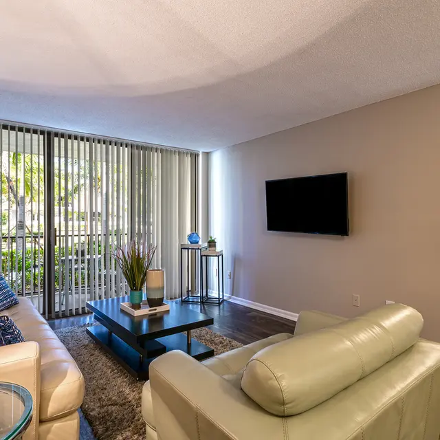 A modern living room featuring a white couch with blue and purple pillows, a soft green chair, and a coffee table. There is a flat-screen TV mounted on the wall and large glass doors leading to a terrace with greenery outside.
