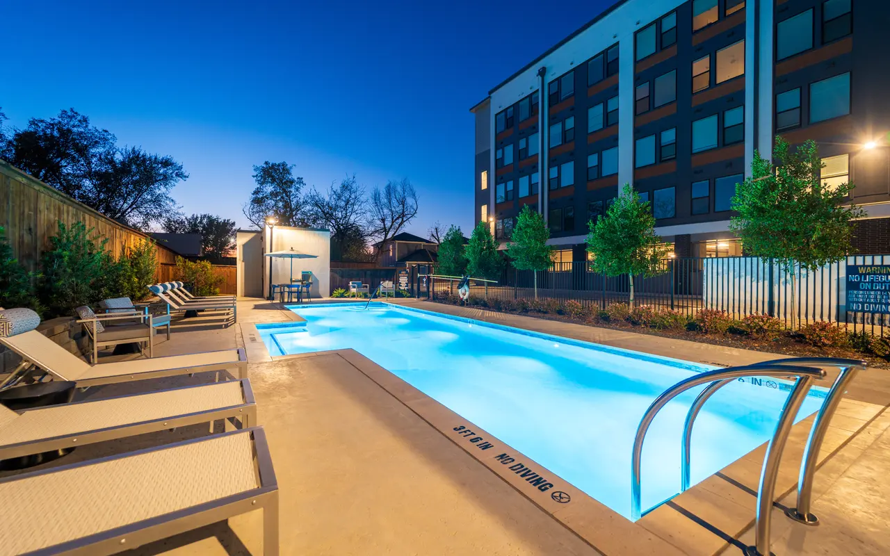A well-lit swimming pool area at dusk with lounge chairs on the side, surrounded by greenery and a modern building in the background.
