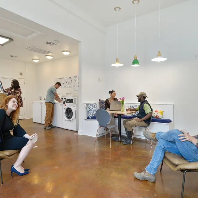 A modern laundry room featuring a couple of people engaged in various activities. On the left, a woman in a black dress and blue heels is sitting in a chair, looking at the camera. In the background, a man is using a washing machine, while another man stands nearby. A group of two people is seated at a table, one wearing a cap and another enjoying a drink.
