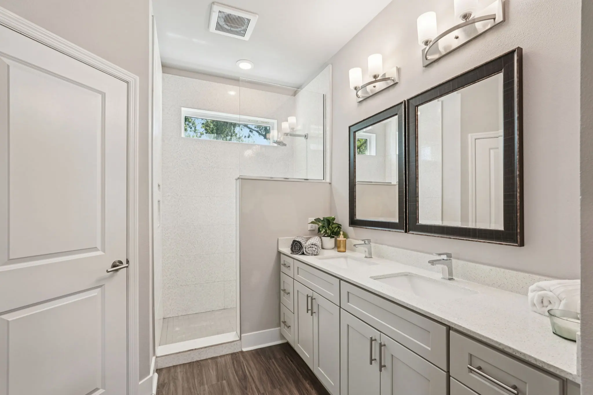 A modern bathroom featuring a spacious shower area, large mirrors, and a sleek vanity with dual sinks. The decor is light and airy with neutral tones and wood-like flooring.