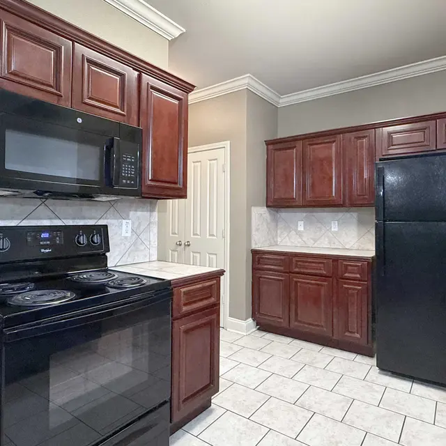 A modern kitchen featuring dark wooden cabinets and black appliances. The kitchen includes a stove, oven, microwave, and refrigerator, with a tiled floor and neutral wall color.