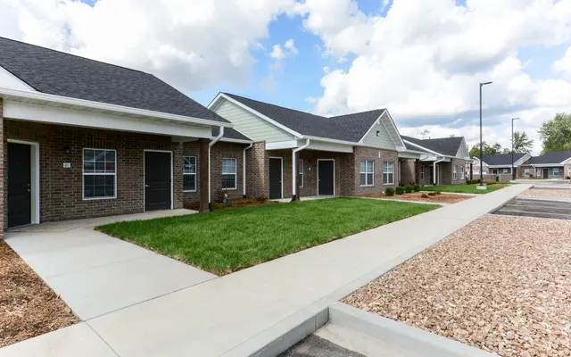 A row of modern single-story brick houses with slanted roofs, set in a manicured landscape featuring concrete sidewalks and patches of grass, under a bright blue sky with scattered clouds.