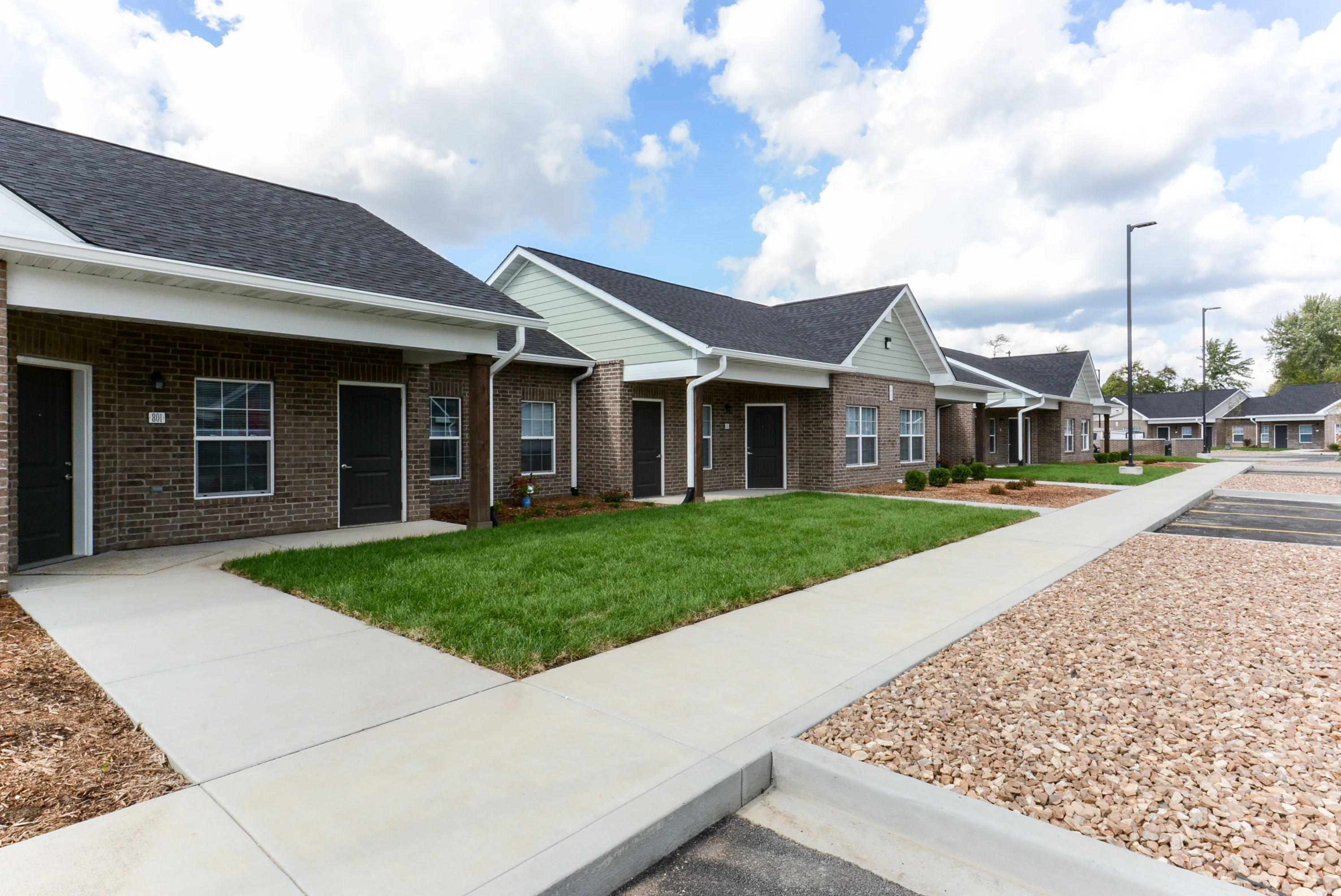 A row of modern single-story brick houses with slanted roofs, set in a manicured landscape featuring concrete sidewalks and patches of grass, under a bright blue sky with scattered clouds.