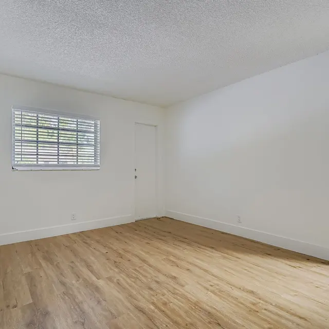 An empty room with light-colored walls and a wooden floor. There is a window with horizontal blinds on one wall and a door on the opposite wall.