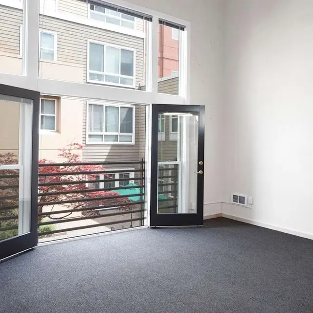 Interior of a bright apartment featuring two open doors leading to a small balcony with a view of neighboring buildings and trees.