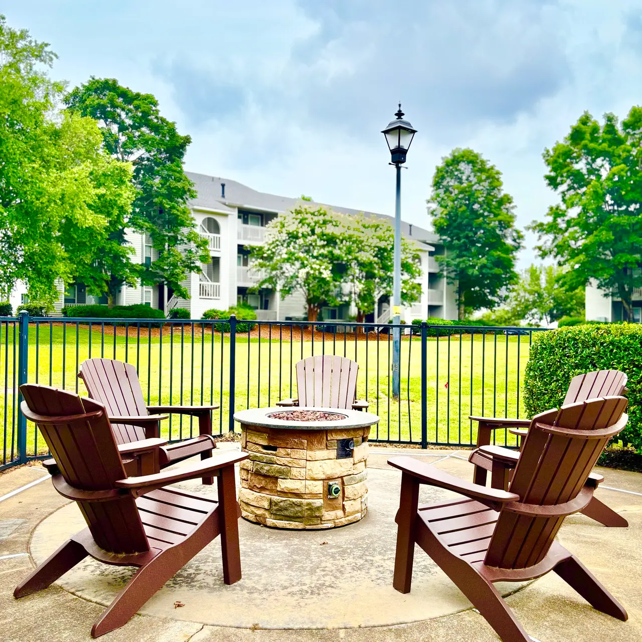 An outdoor fire pit area featuring wooden adirondack chairs arranged around a stone fire pit, with green trees and an apartment building in the background under a cloudy sky.