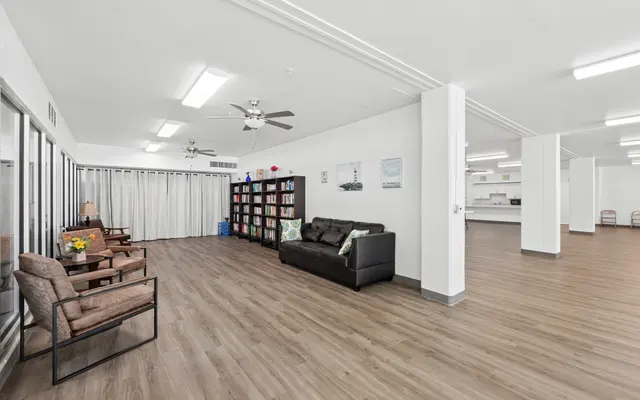 A modern living and communal area featuring a couch, several chairs, a bookshelf, and a kitchen area in the background.