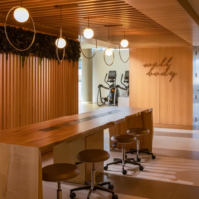 A contemporary interior space featuring a long wooden table with stools, stylish pendant lights overhead, and a wood-paneled wall with plants. The background shows gym equipment, indicating a multifunctional area.