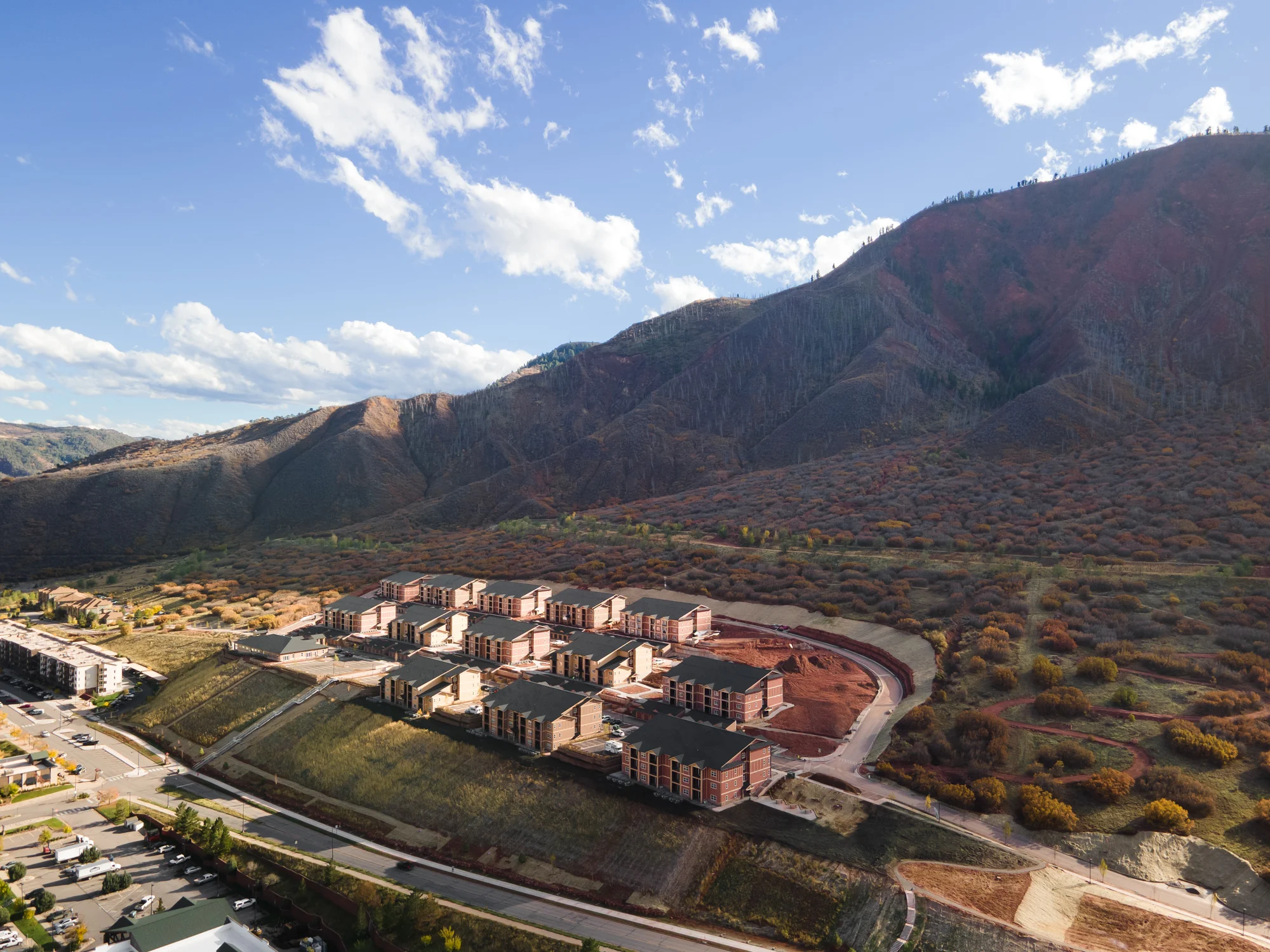 Aerial view of a construction site with multiple buildings on a hillside, surrounded by mountains and trees in autumn colors.