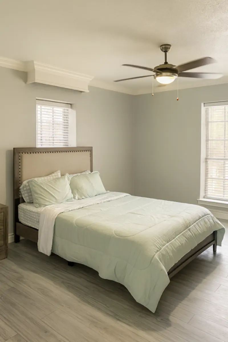 A modern bedroom featuring a bed with a light green quilt and pillows, a ceiling fan, and windows with blinds.