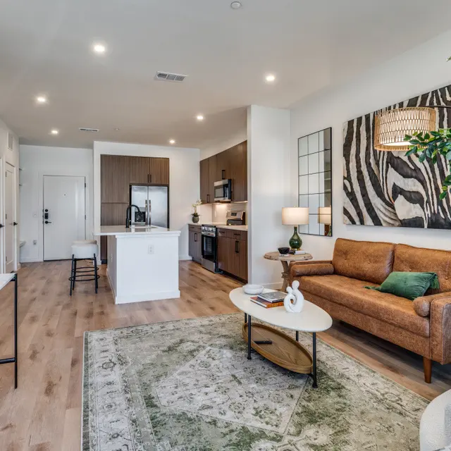 A modern living room featuring a sofa, coffee table, and kitchen area with contemporary appliances. The decor includes plants and artwork.