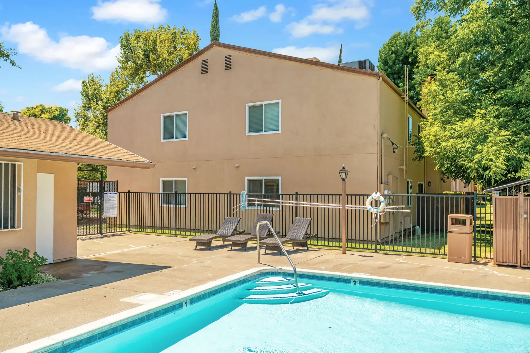 View of a swimming pool area with lounge chairs, surrounded by a fence and an apartment building in the background.