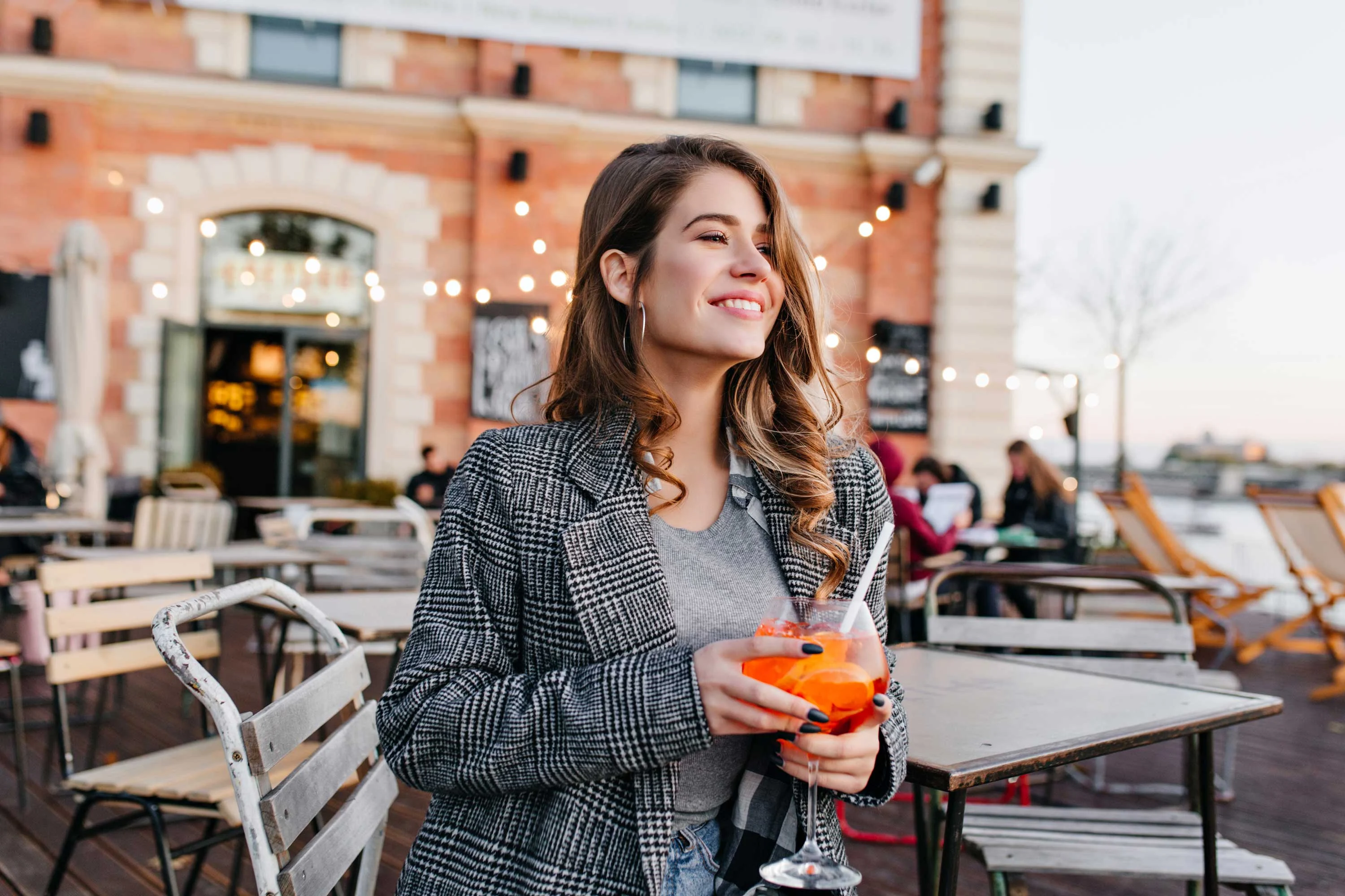 Outdoor Cafe Experience A young woman smiling while holding a cocktail in an outdoor setting with cafe tables in the background.