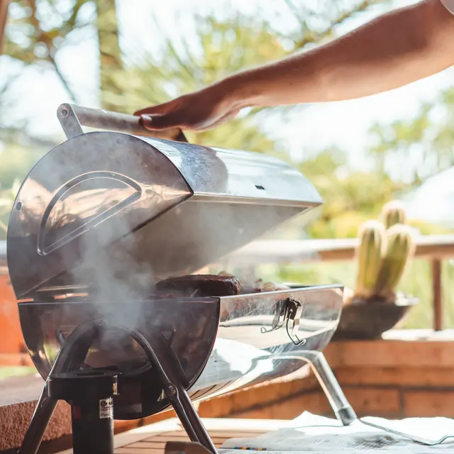Outdoor Grilling Scene A person uses their hand to open a small, shiny grill emitting smoke, set on a table with a bowl in the foreground, and a natural outdoor backdrop with trees and plants.