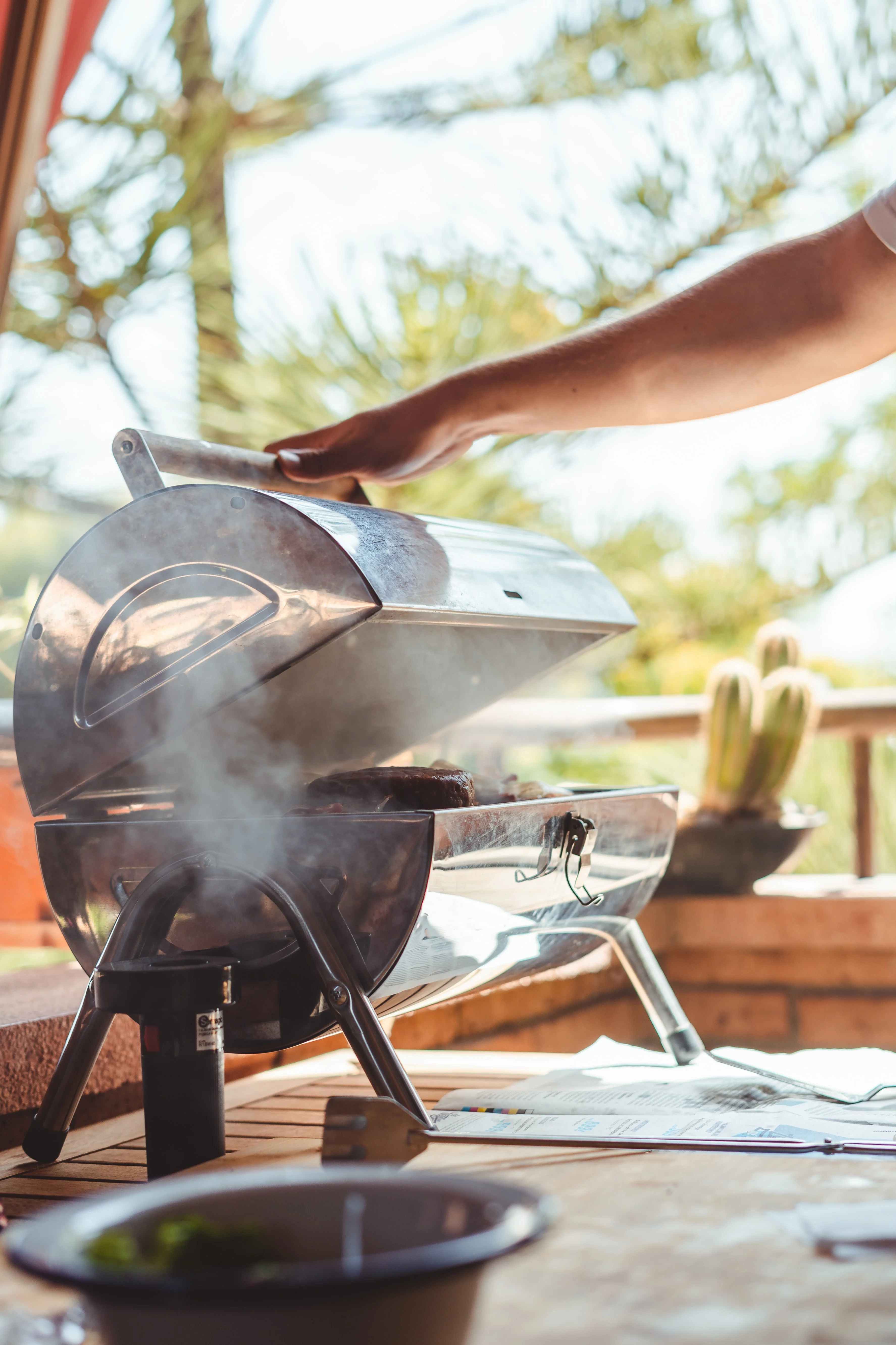 Outdoor Grilling Scene A person uses their hand to open a small, shiny grill emitting smoke, set on a table with a bowl in the foreground, and a natural outdoor backdrop with trees and plants.