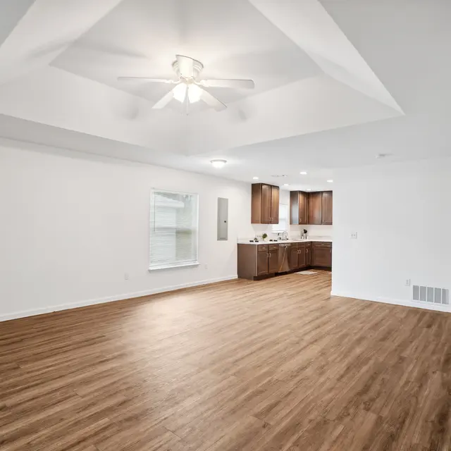 Modern Open Living Space A spacious living area with wooden flooring and a ceiling fan, leading into a kitchen with dark cabinetry.