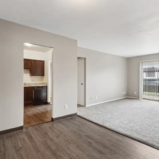 Spacious living room with a light gray carpet and large windows overlooking a backyard. Adjacent kitchen visible through an open doorway.