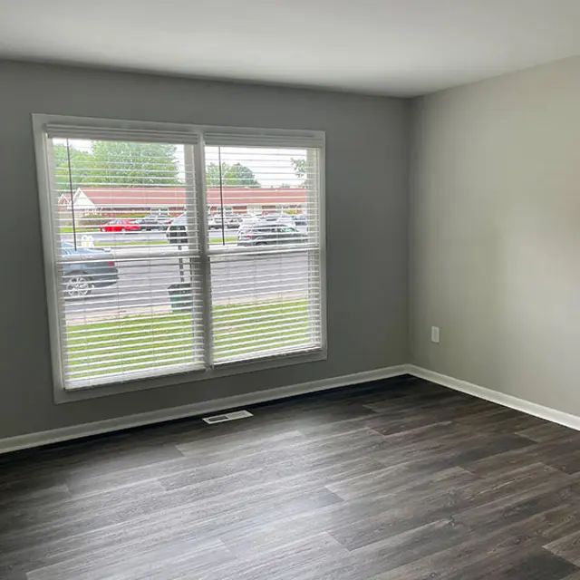 A spacious room with grey walls and dark wooden floor. A large window with blinds is visible on one wall, allowing natural light to enter. The view outside shows a parking area with several cars.