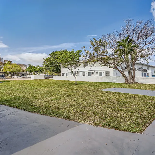 A view of a spacious outdoor area featuring green grass, a tree, and buildings in the background under a clear blue sky.