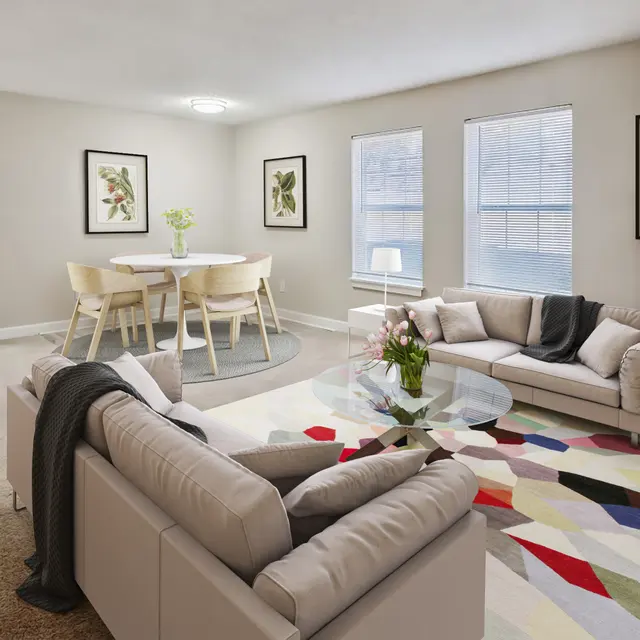 A modern living room featuring two light-colored sofas, a glass coffee table, and a colorful rug. There is a small dining table with chairs, plants on the table, and framed floral art on the walls. The room has large windows allowing natural light to enter.