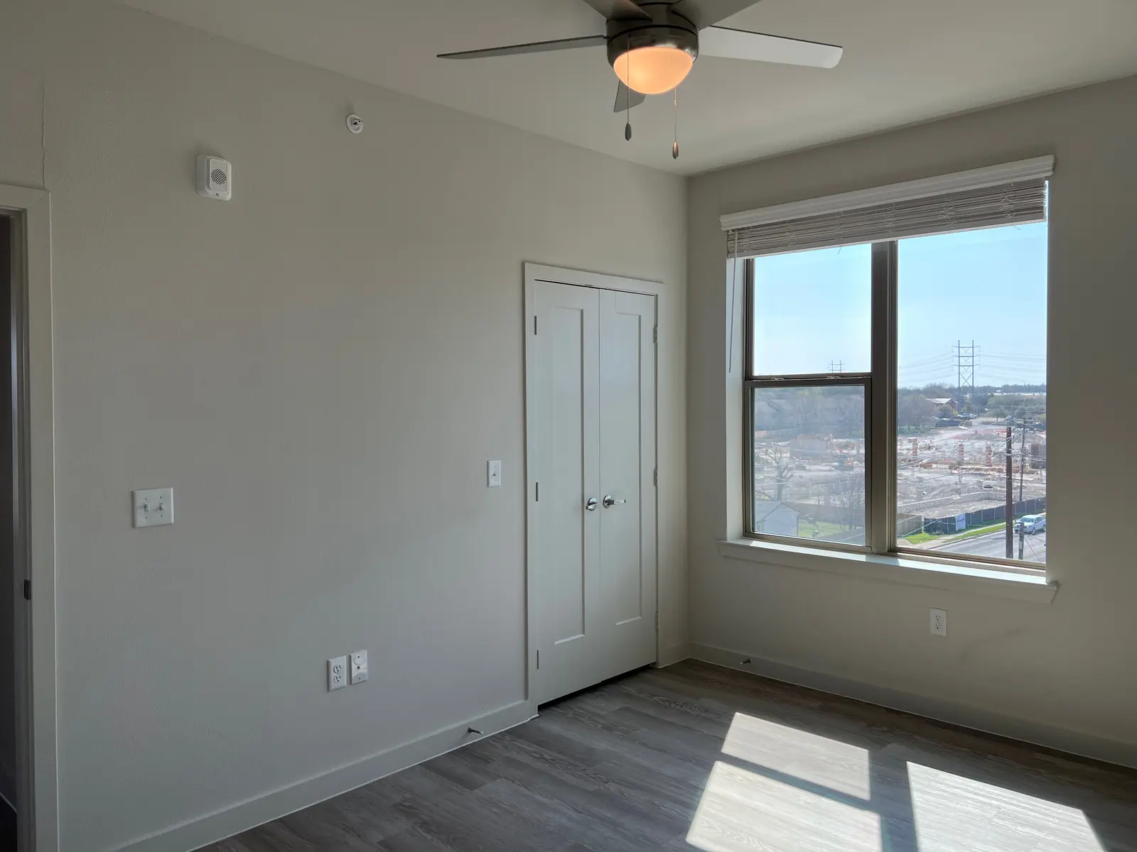 Empty Modern Room with Window and Ceiling Fan A modern, empty room featuring a ceiling fan, large window, and light-colored walls. There is a door on one wall and sunlight streaming in from the window, illuminating the room.