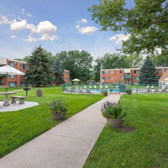 Regency Park Apartments Regency Park - A landscaped apartment complex featuring green lawns, trees, and a swimming pool area with lawn furniture and umbrellas.