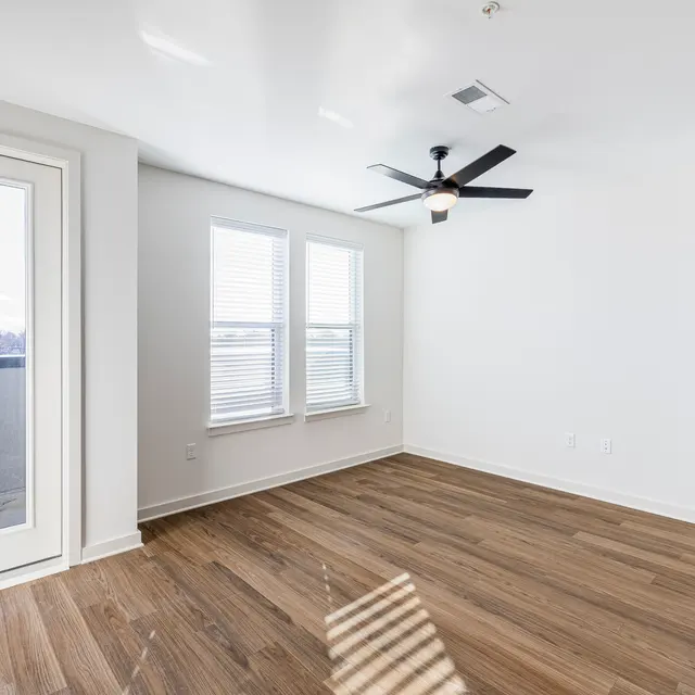 A bright and airy living room with wooden flooring, a ceiling fan, and large windows letting in natural light. There's a glass door leading to a balcony.