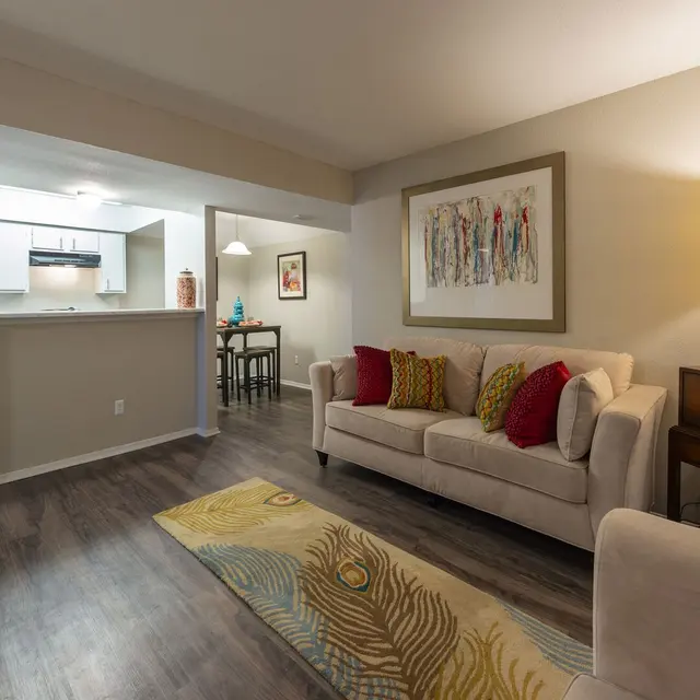 Cozy living room with a light sofa, colorful cushions, and a lamp, leading to an open kitchen and dining area.