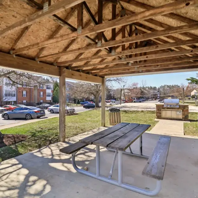 A covered picnic area with a wooden table and benches, surrounded by grass and trees, in a residential area with parked cars in the background.