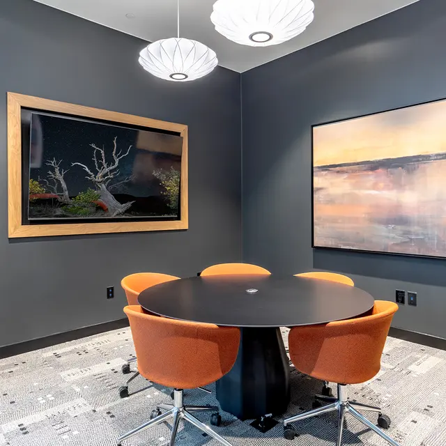 Modern Meeting Room A stylish meeting room featuring a round black table with orange chairs, surrounded by dark gray walls and two large artworks. A potted plant is seen in the corner, and modern pendant lights hang from the ceiling.