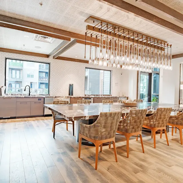 A modern kitchen and dining area featuring a large wooden table surrounded by woven chairs, with a stylish chandelier overhead, and windows providing natural light.