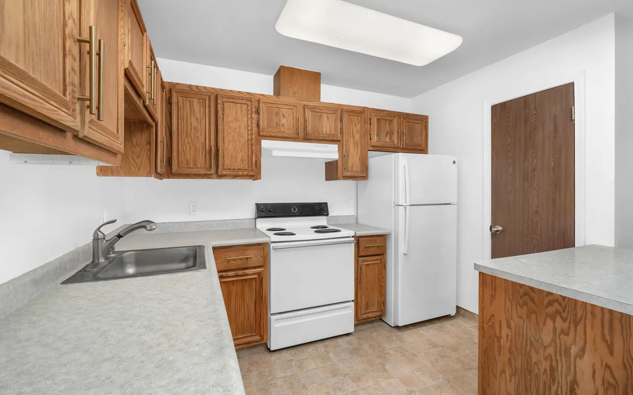 A simple kitchen with wooden cabinets, a white stove, and a refrigerator. The counter is light gray with a stainless steel sink.