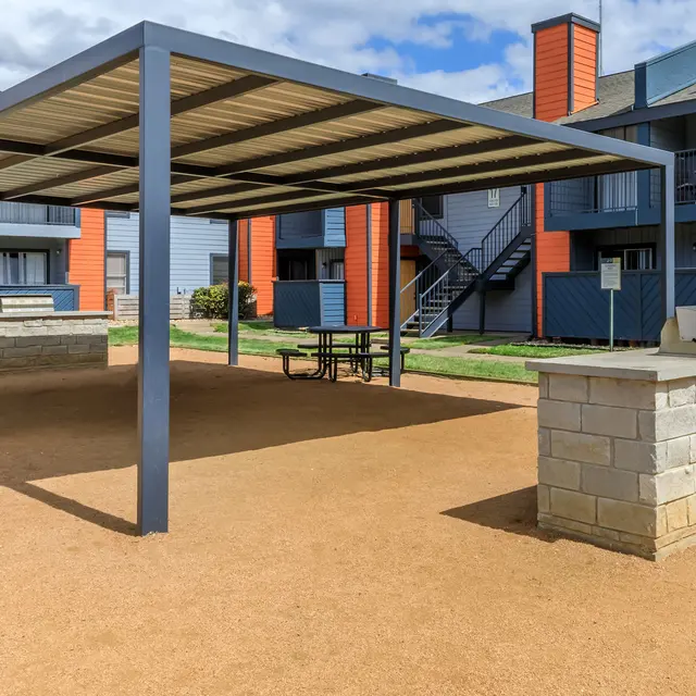 An outdoor communal space in an apartment complex featuring a covered picnic area and a stone grilling station. The backdrop shows buildings with colorful exteriors and staircases.