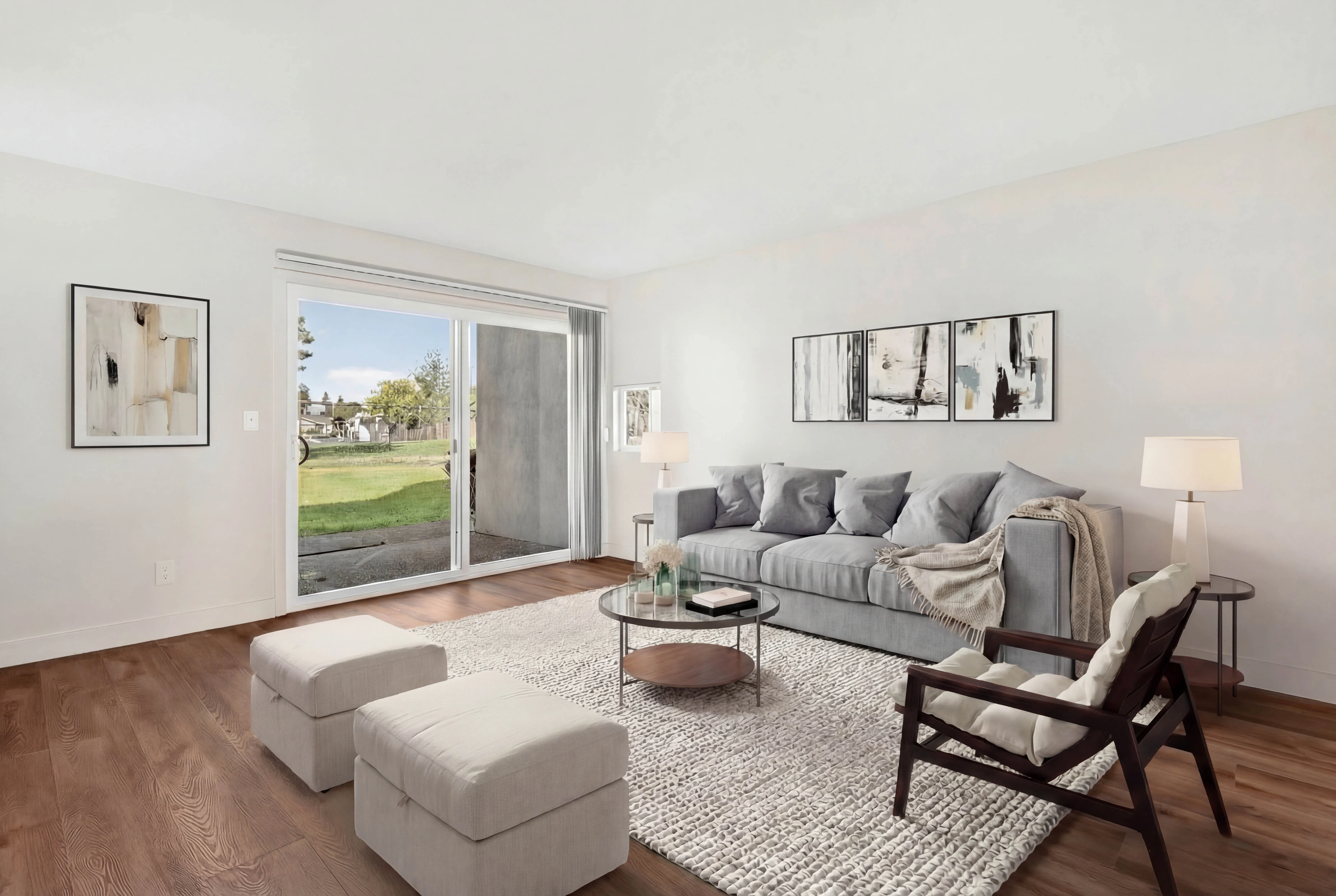 Stylish Modern Living Room A modern living room featuring a light gray sofa with several pillows, a round coffee table, and two ottomans on a textured rug. There is a chair made of dark wood, a lamp, and artwork on the walls. Large sliding doors lead outside, allowing natural light in.