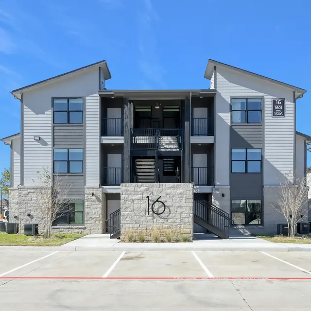 A modern three-story apartment building with a stone base, featuring several windows and balconies. The sky is clear and blue, and there are parking spaces in front.