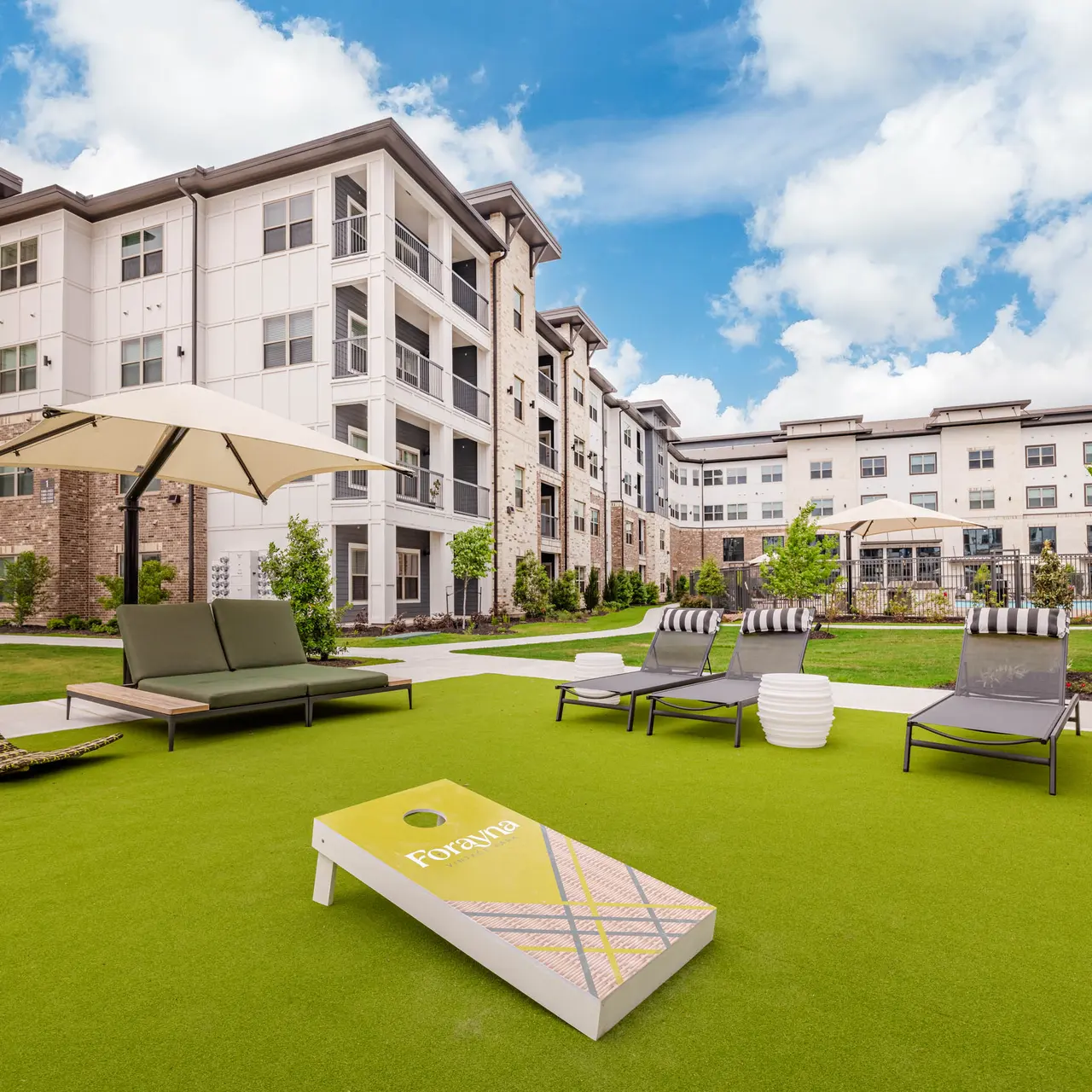 A modern outdoor lounge area at an apartment complex featuring green turf, lounge chairs, an umbrella, and a cornhole game set, with buildings in the background.