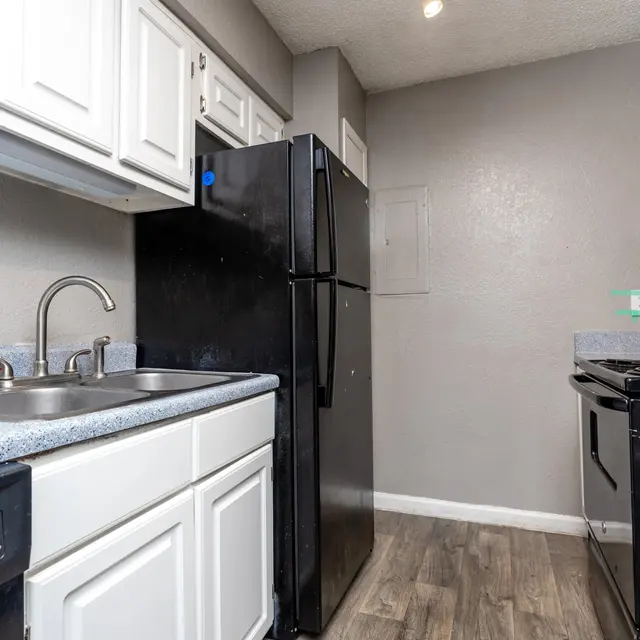 A modern kitchen with grey walls, white cabinets, a black refrigerator, and a black stove. The countertop is grey and there are dual sinks. The flooring is wooden and light-colored.