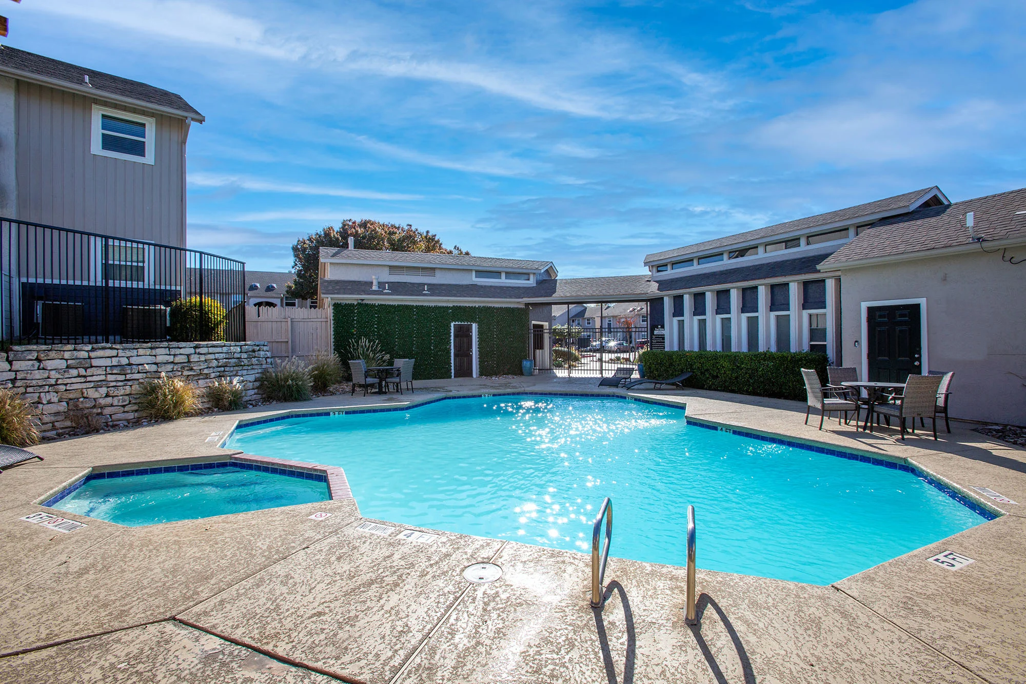 A sparkling blue swimming pool surrounded by a concrete deck, with a few lounge chairs nearby and residential buildings in the background.