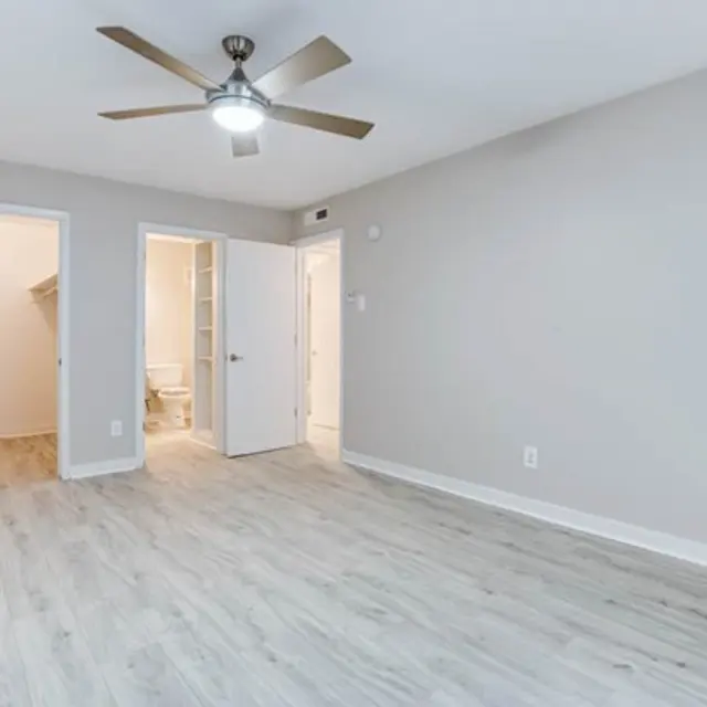 A spacious, empty room featuring light wood flooring, a ceiling fan, and multiple doorways leading to other rooms.