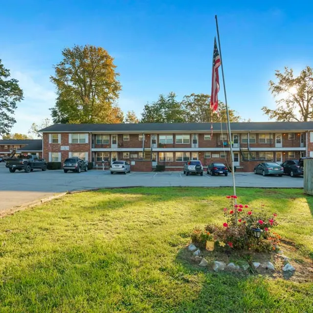 Exterior view of a two-story apartment complex with parked cars and a garden area in the foreground, featuring a flagpole with an American flag.