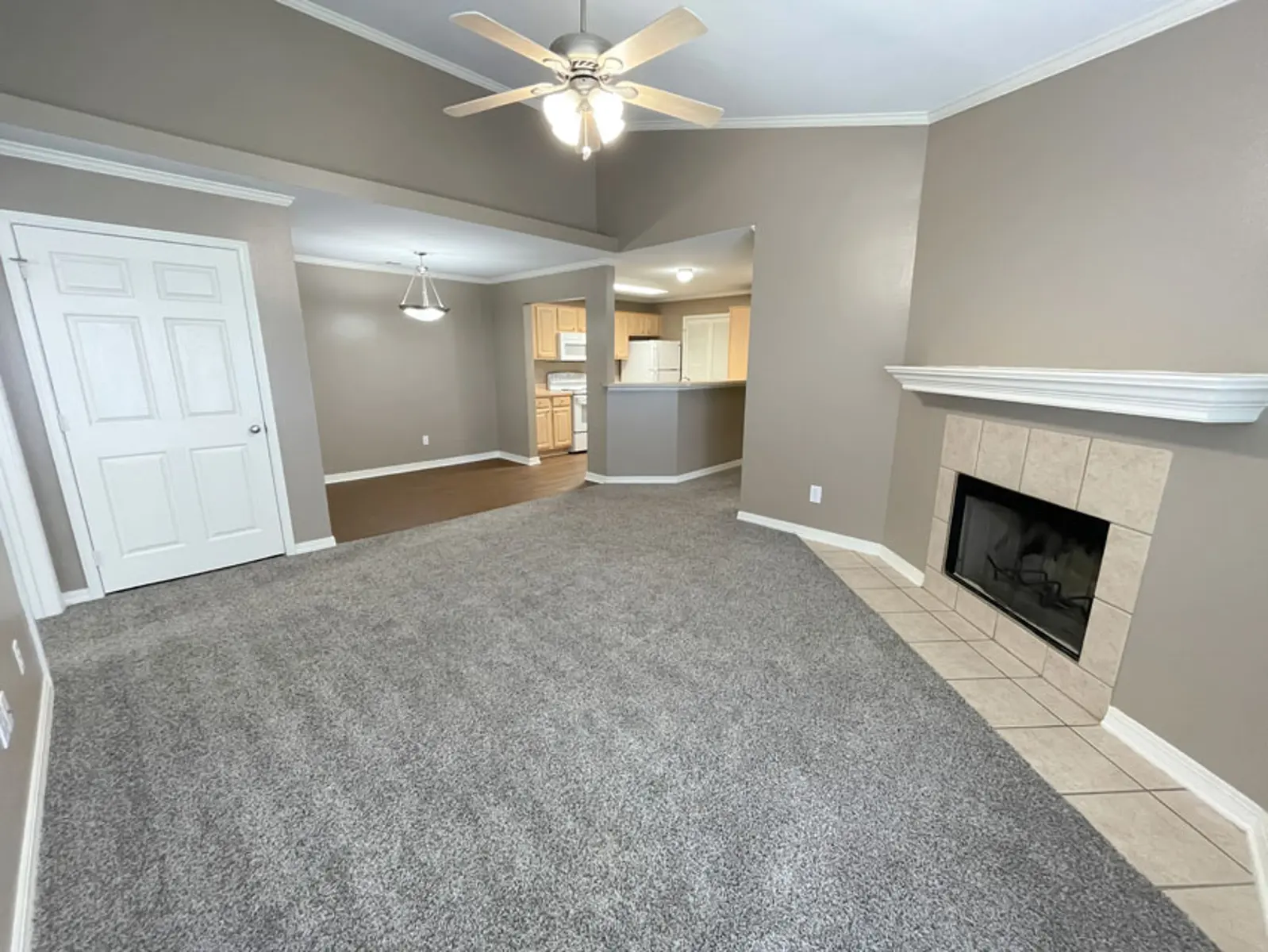 A spacious living room featuring a carpeted floor, a ceiling fan, and a fireplace. The room has beige walls and an open layout connecting to a kitchen area.