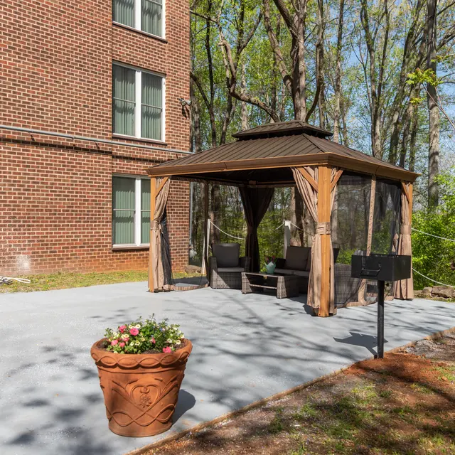 A gazebo with draped curtains over a concrete patio, accompanied by a flower pot, in a green garden area next to a brick building.