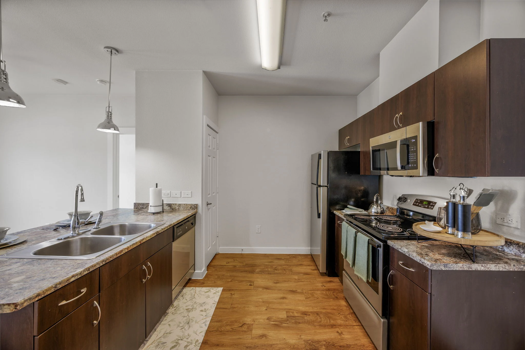 Stylish Modern Kitchen A modern kitchen featuring dark wood cabinetry, stainless steel appliances, and a dual sink. The countertops are made of granite, and the floor consists of wooden planks, creating a warm ambiance. Pendant lights hang from the ceiling, and a large window allows natural light to brighten the space.