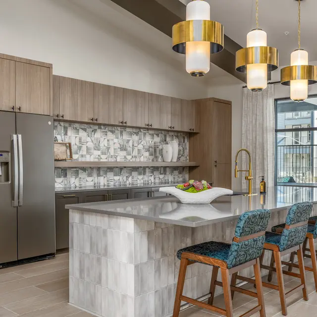 A modern kitchen featuring wooden cabinets, a stainless steel refrigerator, and an island with bar stools. Decorative tile backsplash and large windows provide natural light.