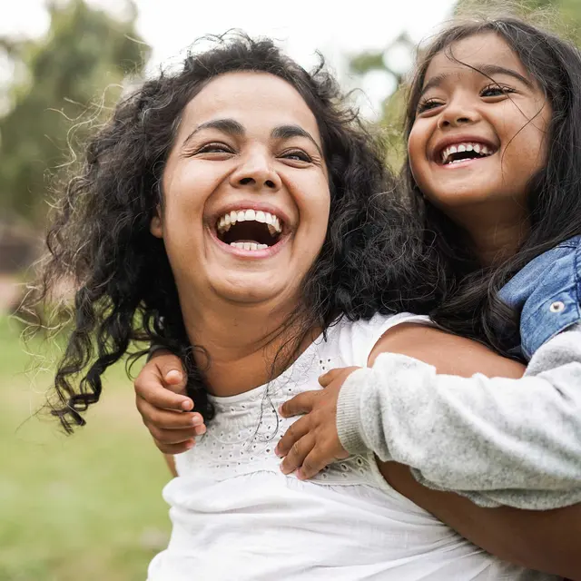 A happy woman carrying a young girl on her back, both laughing joyfully in a lush outdoor setting.